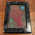 A small black rectangular framed art piece on a wooden table, featuring a red astronaut silhouette with blue stars over a dark striped background.