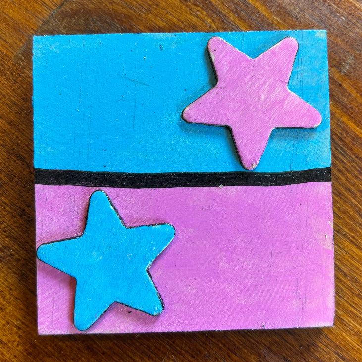 A square handmade craft plaque or magnet featuring a split design of bright blue and pink with two star cutouts, placed on a wooden table.