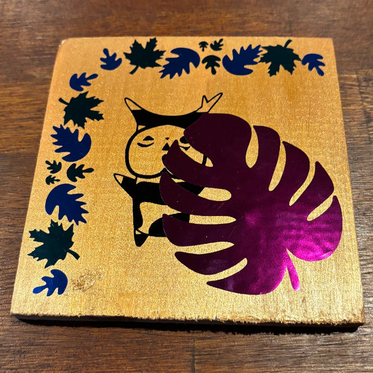 A square wooden coaster with a gold finish, featuring a black silhouette of a cat holding a purple monstera leaf, surrounded by a border of black leaf cutouts on a dark wood background.