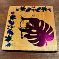 A square wooden coaster with a gold finish, featuring a black silhouette of a cat holding a purple monstera leaf, surrounded by a border of black leaf cutouts on a dark wood background.