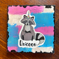 A square wooden plaque or coaster featuring a cartoon raccoon with a unicorn horn and the word 'Unicoon' on a pink, blue, and white painted background, placed on a wooden table.