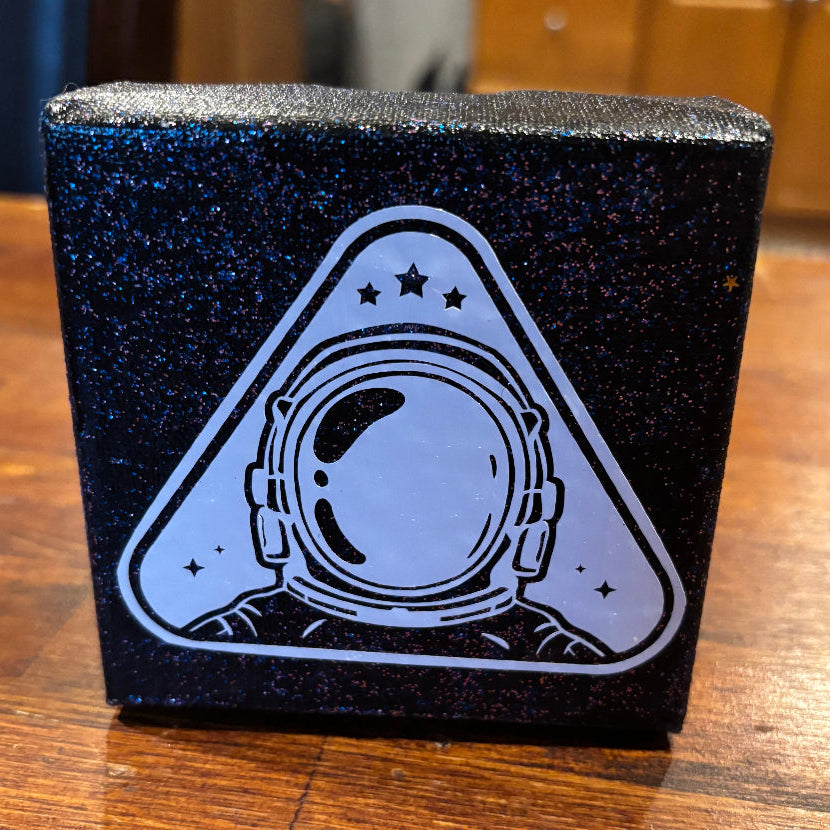 A square black glitter box featuring a white astronaut helmet graphic inside a triangle, sitting on a wooden table.