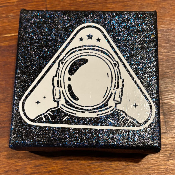 A square black glittery box with a white astronaut helmet graphic on the lid, placed on a brown wooden surface.