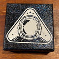 A square black glittery box with a white astronaut helmet graphic on the lid, placed on a brown wooden surface.