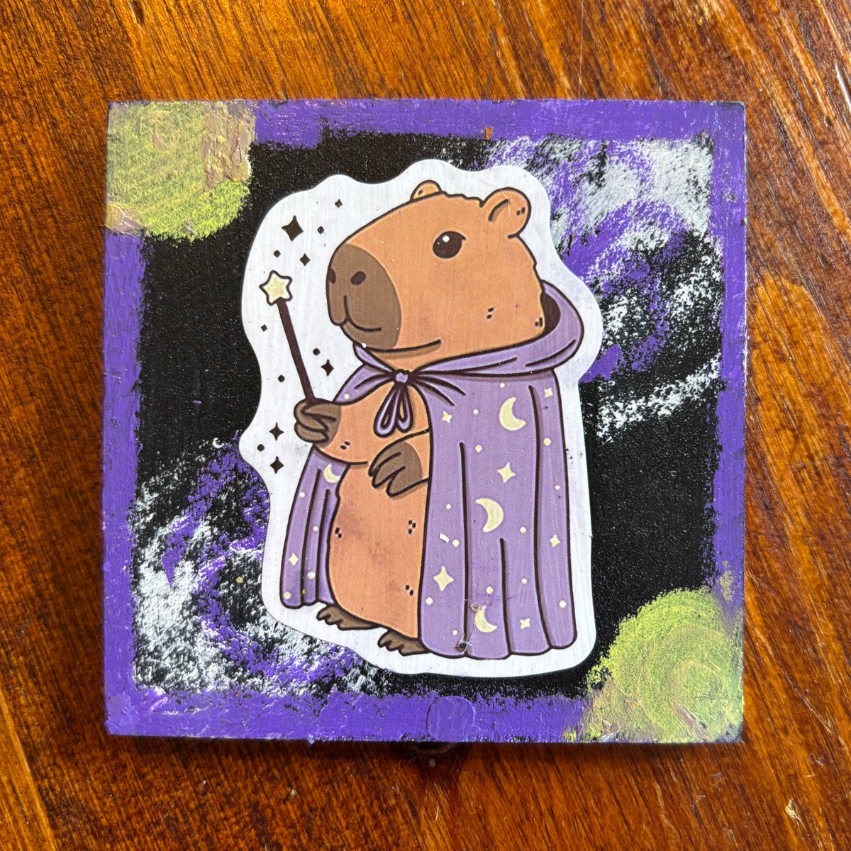 A square handmade art card or sticker featuring a cute capybara wearing a purple wizard robe, set against a black and purple cosmic background with green textured circles, placed on a wooden table.