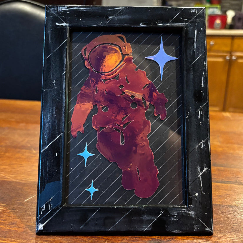 A rectangular black picture frame with a distressed, scratched finish, containing a graphic of an astronaut in red and orange floating against a black space background with blue stars. The frame is displayed standing on a wooden table in a kitchen setting.