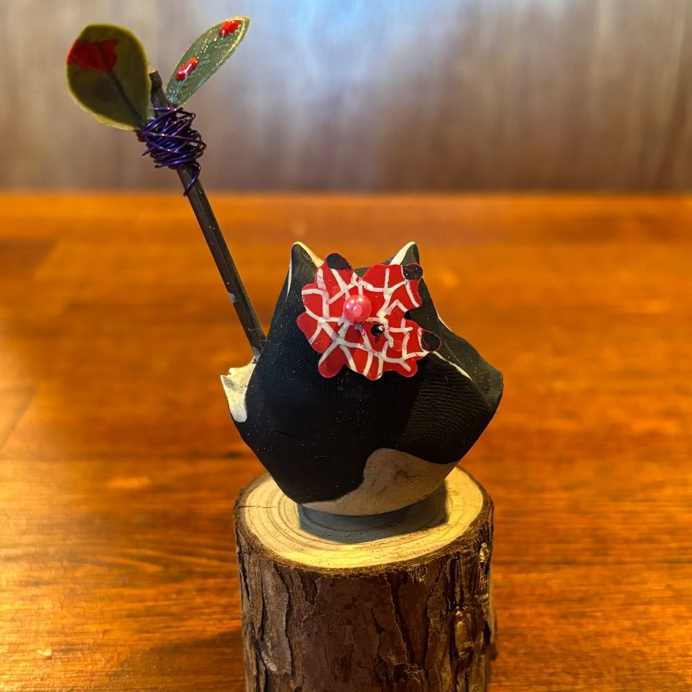 A small black decorative figurine or sculpture with a red and white patterned bow, mounted on a round wood stump base, photographed on a wooden tabletop against a softly blurred brown background.