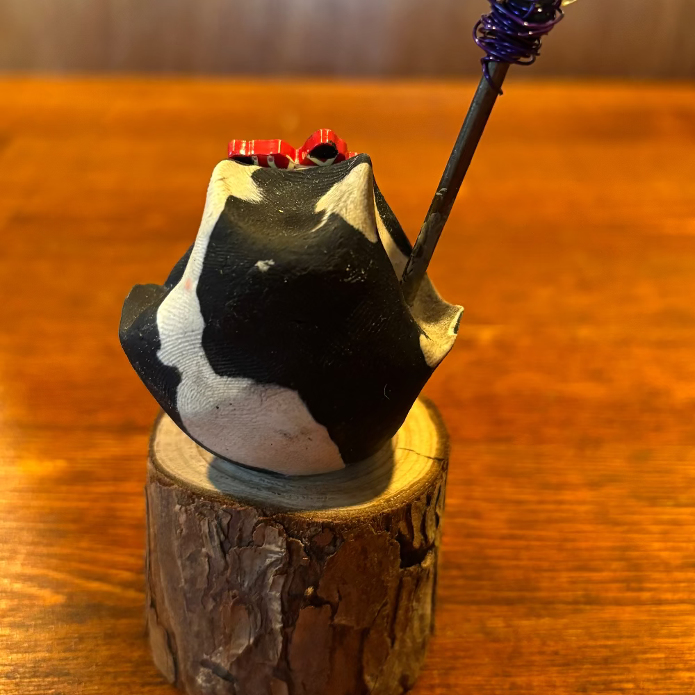 A small black and white ceramic bird figurine with red accents, perched on a rustic wooden log block. A decorative twig branch with green leaves and a red berry hangs from its beak.