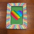 A square decorative picture frame made from many colorful pencils arranged in a radial pattern around a white distressed border, holding a green card with a rainbow-striped heart design, photographed on a wooden tabletop.