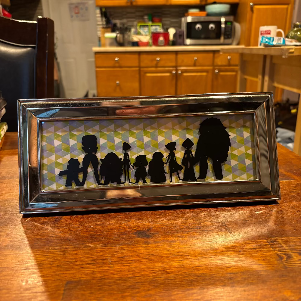 A rectangular framed silhouette art piece featuring a family of seven in black silhouette against a green, yellow, and white geometric patterned background, displayed on a wooden table in a kitchen setting.