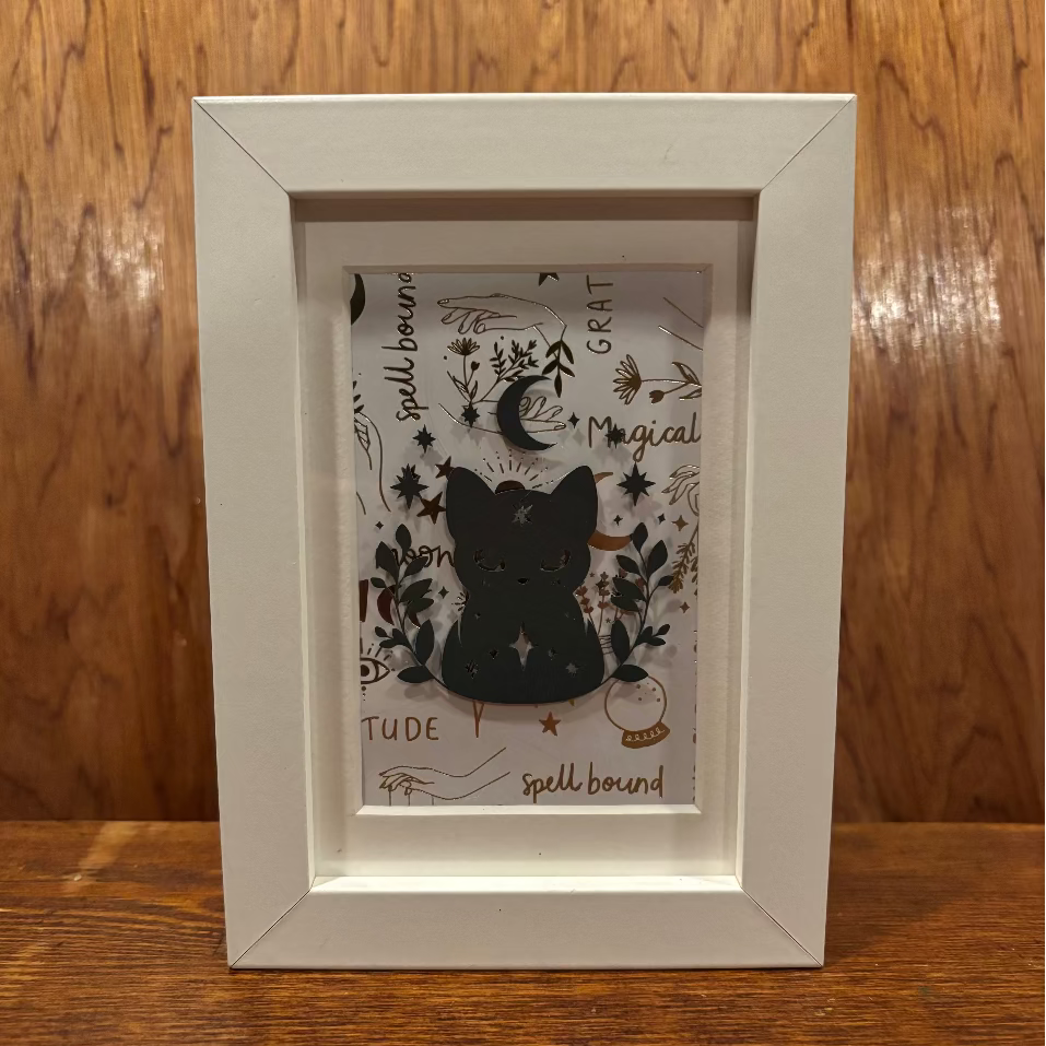 A white square framed art print featuring a black cat silhouette surrounded by celestial and mystical illustrations, set against a warm wood background.