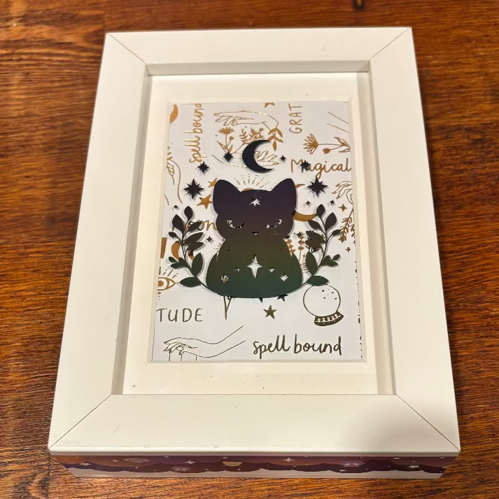 A square white-framed art print featuring a dark green cat silhouette surrounded by celestial and spell-themed illustrations, displayed on a wooden tabletop.