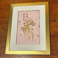 A framed art print featuring a gold celestial design of a crescent moon and stars on a light pink background, housed in a white mat and a gold frame, placed on a wooden surface.