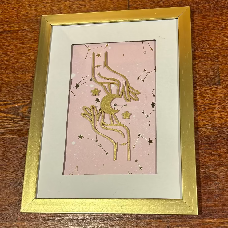 A framed art print featuring a gold celestial design of a crescent moon and stars on a light pink background, housed in a white mat and a gold frame, placed on a wooden surface.