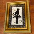 A framed art print featuring a black silhouette of a cat sitting on a mushroom, set against a white background with black and gold botanical details. The print is housed in an ornate gold frame and displayed on a dark wood surface.