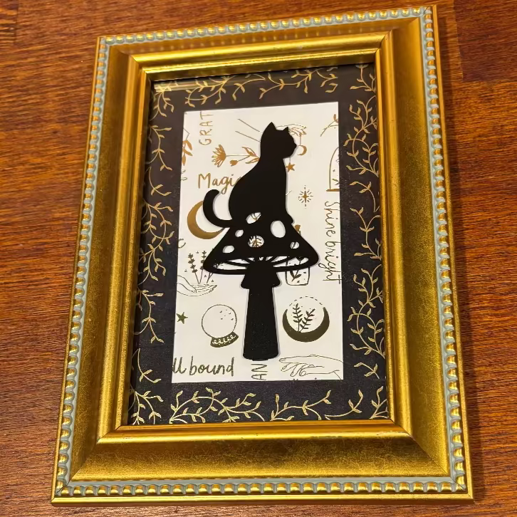 A framed art print featuring a black silhouette of a cat sitting on a mushroom, set against a white background with black and gold botanical details. The print is housed in an ornate gold frame and displayed on a dark wood surface.