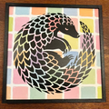 A framed square art print featuring a stylized black pangolin surrounded by pastel multicolor blocks, displayed on a dark wood surface.
