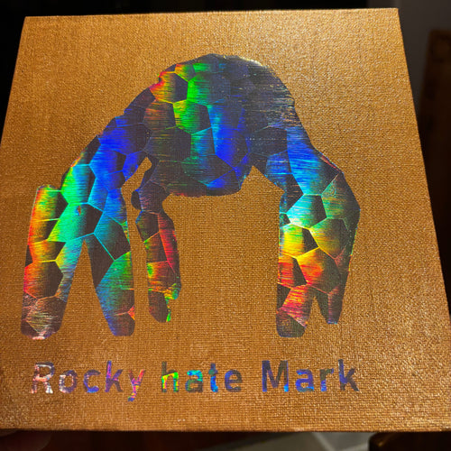 A square gold canvas print featuring a large, colorful geometric handprint in an iridescent holographic finish, with the text 'Rocky hate Mark' printed vertically on the right side.